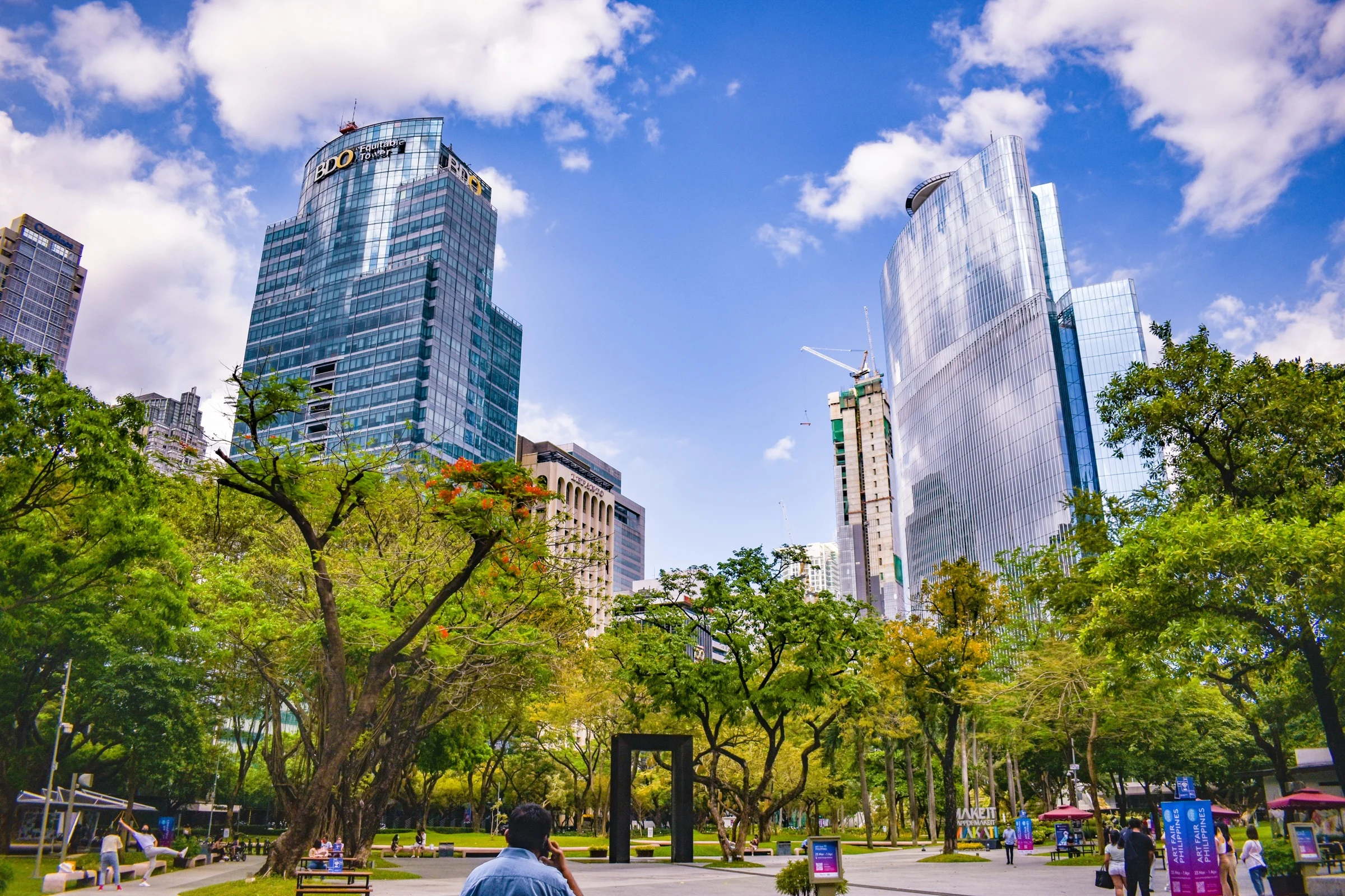 Makati’s towers rise above a green city park, with people walking and gathering beneath the trees.